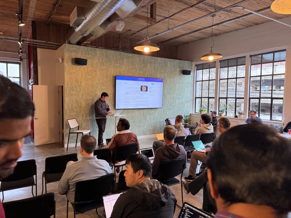 A photograph captures a technical presentation or meeting taking place in a loft-style office space, showing a speaker standing next to a large display screen while an audience of mostly men sits and watches, many with open laptops.