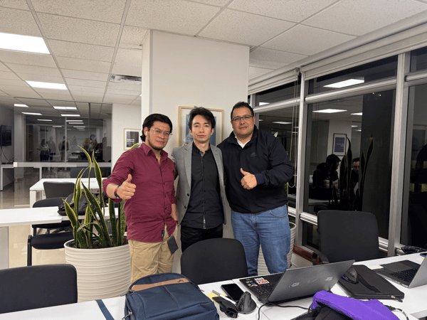 A group photo capturing three men standing together in a modern, brightly lit office space, posing behind a desk that holds several open laptops and bags. Two of the men are giving a thumbs-up gesture.