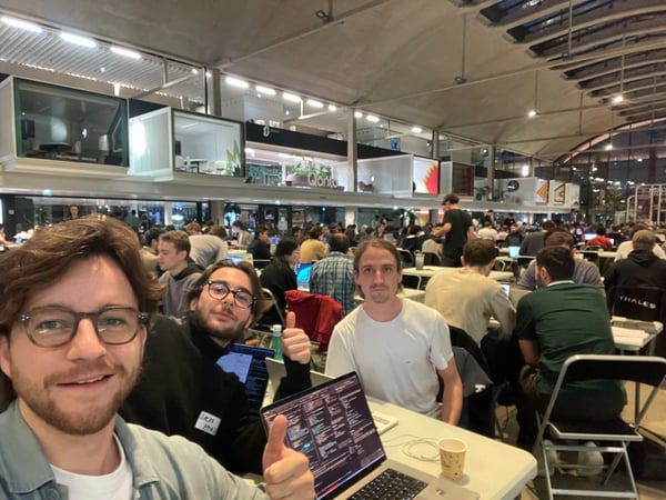 A group selfie of three young men smiling and giving thumbs-up while sitting at a table with open laptops displaying code, set against a backdrop of a crowded indoor event, likely a hackathon or large coding workshop.