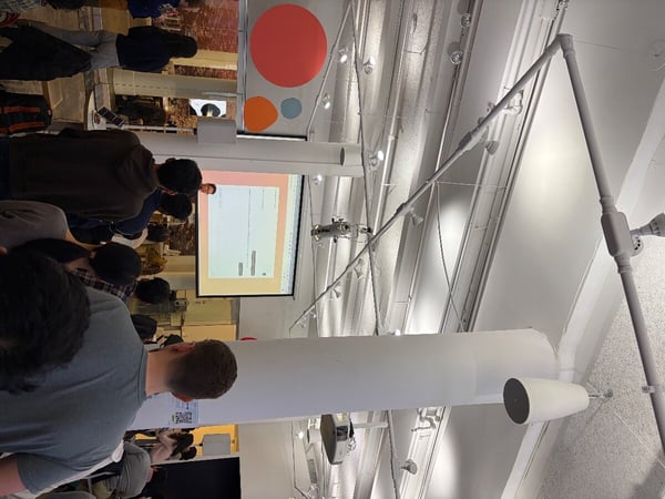 A photograph showing an audience from behind as they watch a presentation on a projector screen in an indoor space with high ceilings and exposed pipes. The image is oriented sideways (rotated 90 degrees counter-clockwise).