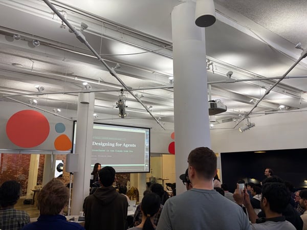 A group of people are gathered in a modern office space for a presentation titled 'Designing for Agents'. A woman stands at the front by a large screen while the audience watches from their seats.