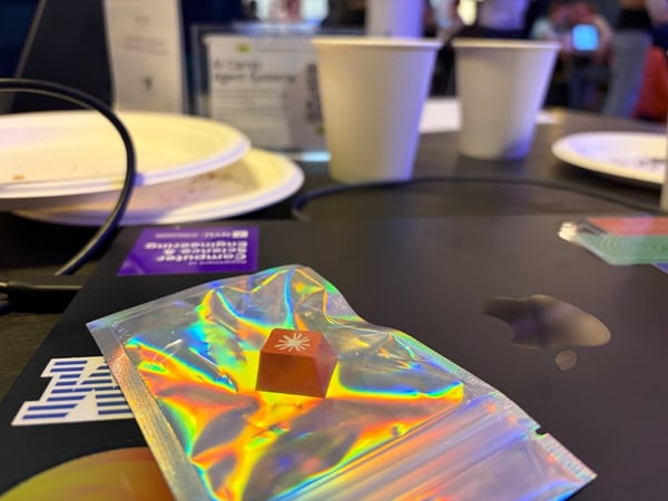 A close-up photograph of an orange mechanical keyboard keycap with a white starburst icon, resting on a holographic silver pouch atop a laptop.