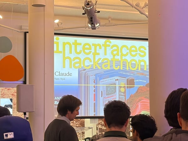 A group of people are attending an event where a large screen displays 'interfaces hackathon' and 'Claude'. The scene appears to be an indoor presentation or workshop setting.
