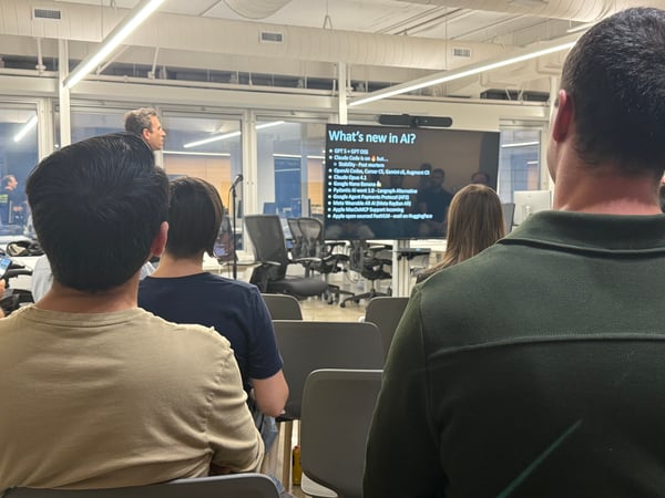 A photograph capturing an audience attending a technical presentation in a modern office space, viewed from behind the attendees, with the screen displaying a bulleted list titled "What's new in AI?".