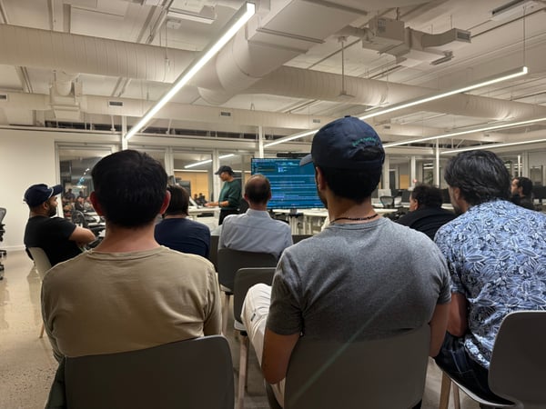 A candid photograph taken from the back of an audience attending a technical presentation in a modern office space with exposed ceilings.