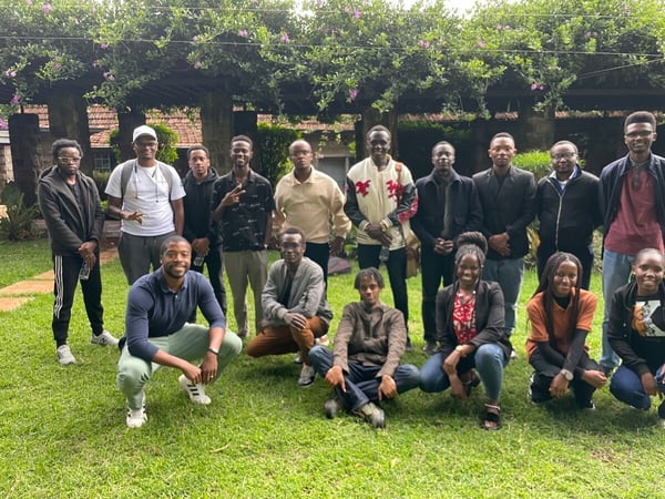 A group of sixteen young people, mostly men, are posing for a group photograph outdoors on a green lawn in front of a stone building covered with flowering vines.