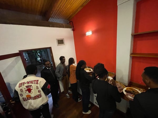 A group of people are lined up indoors, seemingly at a buffet or food service area, with several individuals holding plates of food.
