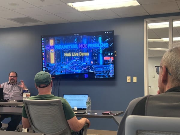 A man is giving a presentation in an office setting, with a large screen behind him displaying 'MOE PARAMETERS, MOE PROBLEMS' and 'MoE Live Demo'.