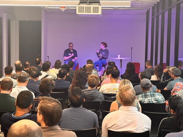 A photograph taken from the perspective of the audience, showing a large crowd attending an indoor fireside chat or presentation featuring two speakers seated on stools on a stage lit with purple ambient light.