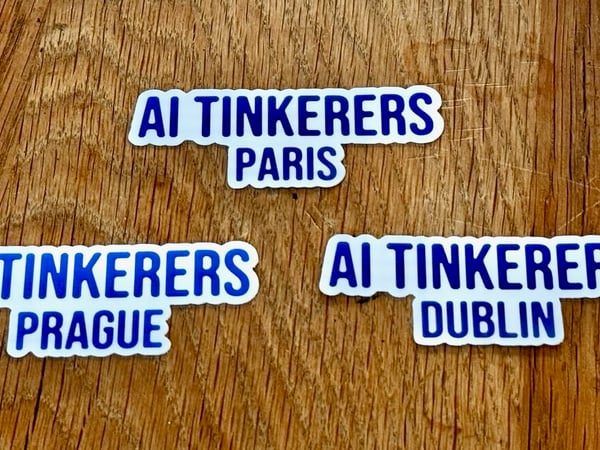 A photograph shows three identical promotional stickers for "AI Tinkerers" chapters (Paris, Prague, and Dublin) arranged on a textured wooden surface. The stickers feature bold blue text on a white background.