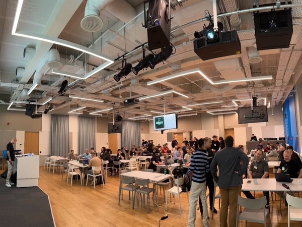 A wide-angle photograph capturing a large indoor event or workshop in a modern conference hall, showing numerous attendees seated at white tables and a presentation screen on the left.