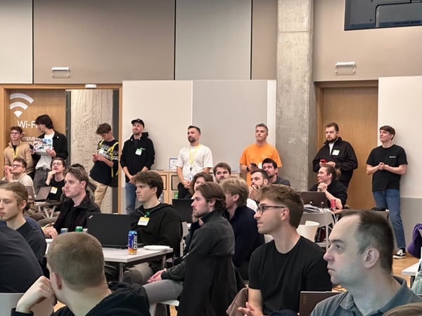 A candid photograph captures a large group of attendees, primarily young men, gathered in a modern indoor venue, likely attending a presentation or workshop. Many people are seated at tables with laptops open, while others stand along the perimeter, all generally facing the front of the room (out of frame).