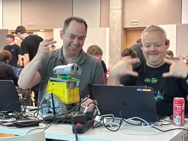 Two men enthusiastically pose with a custom-built, WALL-E-like robot prototype and their laptops, surrounded by a tangle of cables and power strips at an indoor technical event or hackathon.