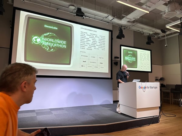 A photograph capturing an indoor event presentation, showing a speaker standing behind a podium in front of a large screen displaying details for the 'ElevenLabs Worldwide Hackathon' and a partial view of an audience member in the foreground.