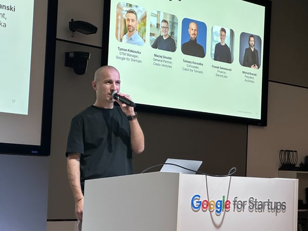 A bald man wearing a black t-shirt is speaking into a microphone behind a white podium branded with the "Google for Startups" logo during an indoor presentation or conference.