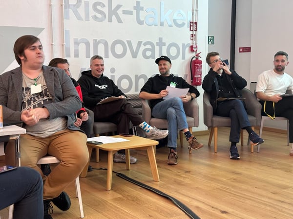 A candid photograph captures a group of six men seated in armchairs during an indoor meeting or workshop, with motivational text reading 'Risk-taker Innovator Vision' visible on the wall behind them.