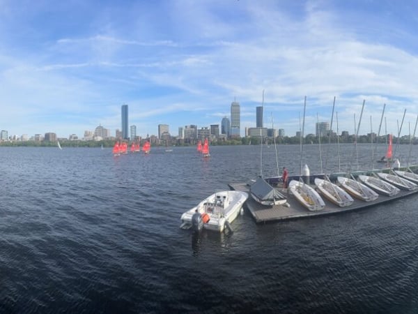 A panoramic photograph of a wide river featuring sailboat docks and a city skyline in the background under a blue sky.
