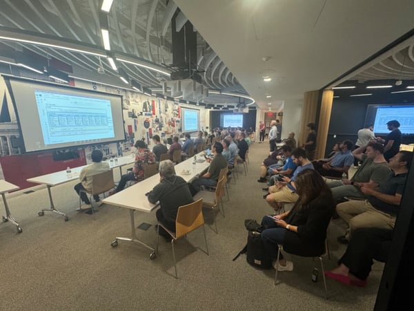 An indoor photograph of an audience seated in a modern lecture hall, watching a technical presentation on a large projector screen.
