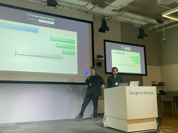 A photograph captures two men presenting on a stage, standing near a podium labeled "Google for Startups." The main projection screen behind them displays a software interface titled "Cockpit Safety Monitor," simulating a chat interface.