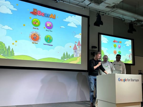 A photograph capturing three men giving a presentation on stage, showcasing a children's educational application called 'BearAI' on two large projection screens behind them.
