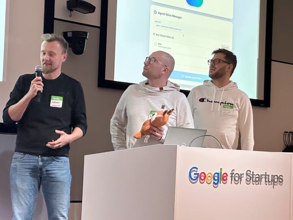Three men are giving a presentation or demonstration, with one man speaking into a microphone, in front of a large screen displaying a software interface labeled "Agent Voice Manager." The podium in front of them is branded "Google for Startups."