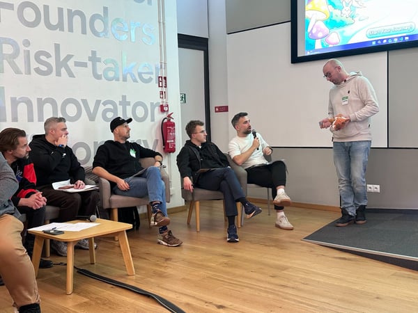 A photo showing an indoor panel discussion or presentation where a standing man addresses five seated men in a modern event space. The background features large silver text on a white wall and a screen displaying colorful game-like graphics.