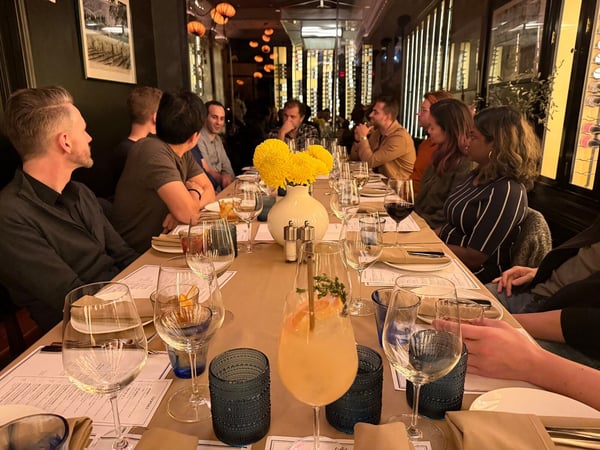 A candid photograph captures a group of ten people seated at a long, formally set dining table in a dimly lit restaurant, engaged in conversation during a meal. The foreground features several glasses, menus, and a cocktail garnished with grapefruit and herbs, alongside a centerpiece of bright yellow flowers.