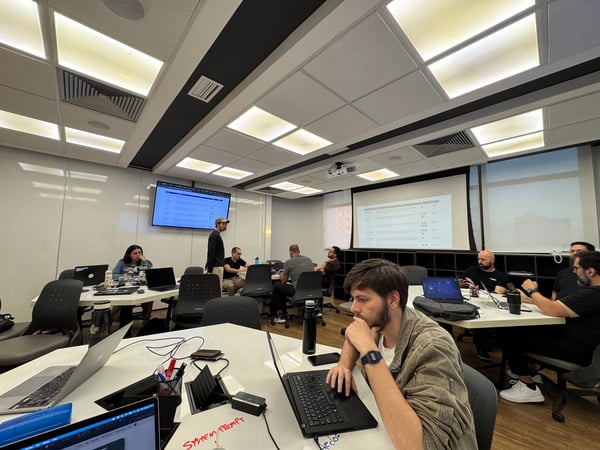 A group of people are working and collaborating in a modern office or conference room equipped with laptops and large display screens.