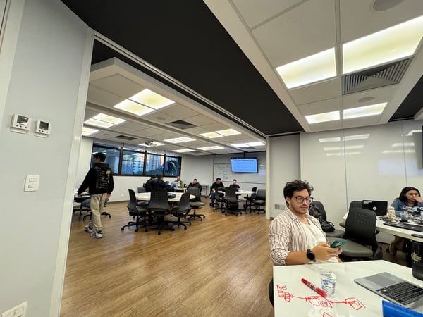 A group of people are working in a modern office or coworking space featuring whiteboard walls, circular tables, and laptops.