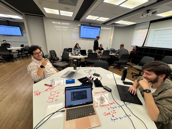 A group of people are working and collaborating in a modern office or coworking space, with diagrams and notes written directly on one of the white tables.