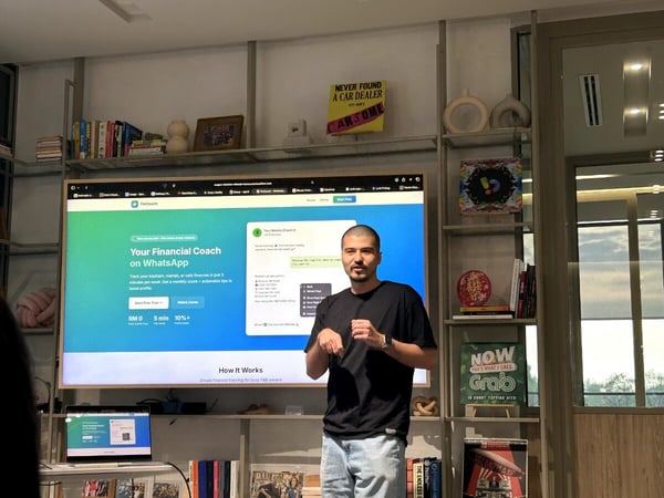 A man in a black t-shirt and light-colored jeans stands in front of a large screen presenting a financial coaching service called 'FinCoach'.