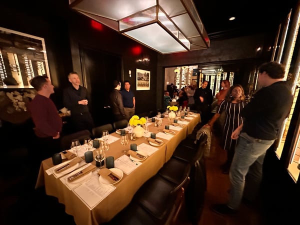 A candid photo captures a group of people socializing around a long dinner table set with place settings and yellow flowers in a dimly lit, upscale private dining room with dark walls and modern lighting fixtures.