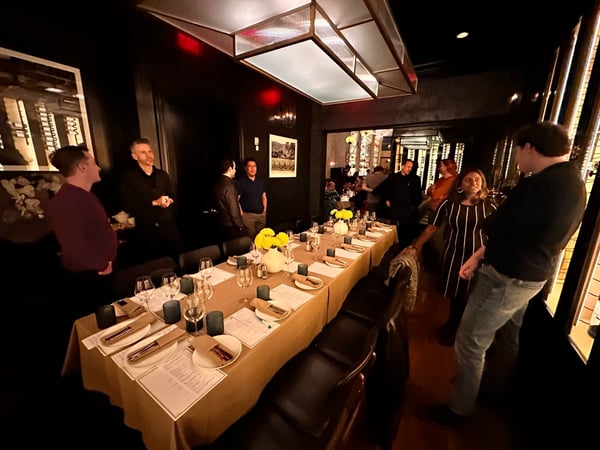 A low-light photograph captures a social gathering in a private dining room, featuring a long table set with place settings, wine glasses, and bright yellow floral centerpieces.