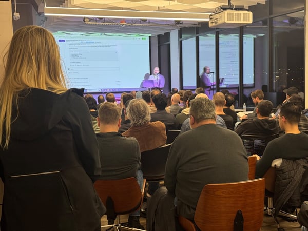 A photograph captures a large audience seated in a dimly lit room attending a technical presentation, with a speaker visible on a stage in front of a brightly illuminated projector screen displaying code or technical content. The image is taken from the back of the room, focusing on the backs of the attendees' heads.