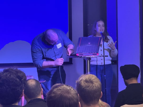 A photograph capturing an indoor technical presentation or demo, where a woman speaks into a microphone while a man bends over to connect a cable to a laptop on a stand, with an audience visible in the foreground.