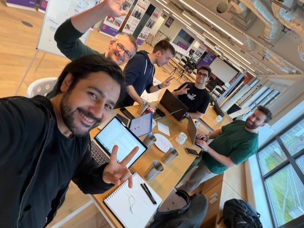 A high-angle group selfie shows five men gathered around a wooden table in a brightly lit modern office, many of whom are smiling and making peace signs or waving. Laptops and notebooks are scattered across the table, suggesting a collaborative work session.