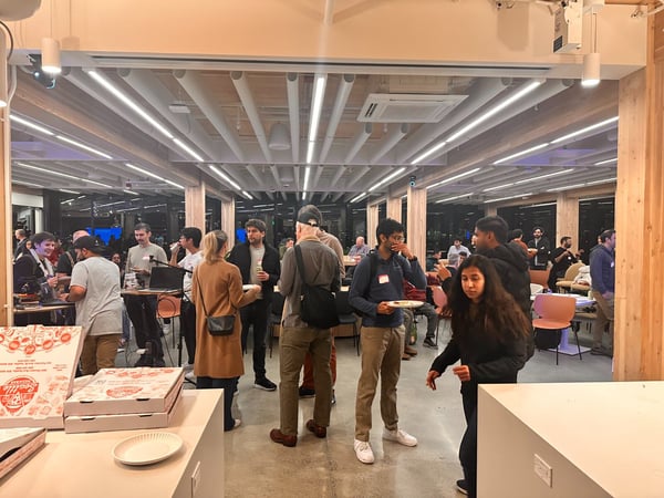 A candid photograph capturing a social or networking event indoors, showing a large group of people mingling, holding plates, and eating near a table stacked with pizza boxes in a modern office space.