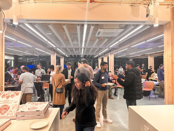 A candid photograph capturing a bustling indoor social or networking event, showing a large group of people conversing and eating (with pizza boxes visible in the foreground) in a modern space featuring wood beams and strip lighting.