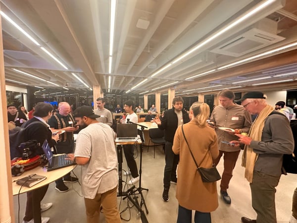 A candid photograph captures a group of people mingling and networking at an indoor evening event, characterized by modern architecture, linear lighting, and several laptops set up for use.