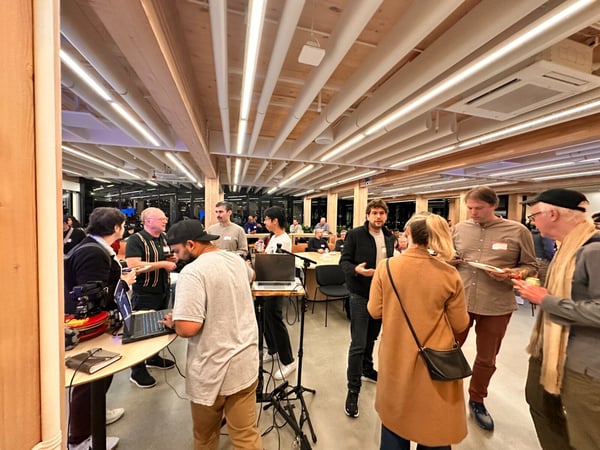 A candid photo captures a group of people mingling and discussing at an indoor social or professional event, featuring modern architecture with wood accents and bright linear ceiling lights.
