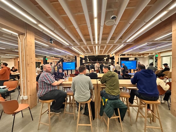 A candid photograph taken indoors showing a large group of people gathered in a modern space featuring exposed wooden beams and columns, high tables, and concrete floors. Several individuals are seated at a long table in the foreground, while others are socializing near large blue screens in the background.