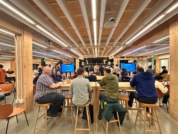 A candid photo capturing a large group of people gathered in a modern, well-lit indoor space featuring high wooden ceilings and beams, sitting at tall tables and socializing.