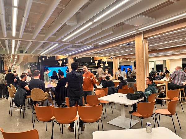 A candid photograph capturing a large group of people socializing and mingling in a modern, brightly lit indoor space, characterized by high ceilings, wooden beams, and contrasting orange chairs.