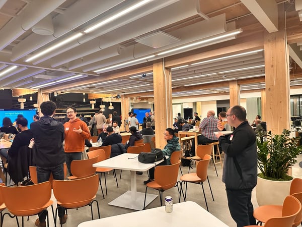 A candid photograph capturing a large group of people socializing and networking in a modern, brightly lit indoor event space featuring exposed wooden beams, white tables, and distinctive orange chairs.