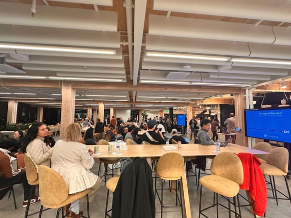 A candid photograph captures a large group of people gathered indoors in a modern, brightly lit communal space featuring exposed wooden beams and long high tables with tan bar stools.
