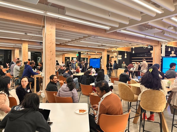A large group of people is gathered indoors in a spacious, modern venue characterized by wooden architectural beams and columns, likely attending a social or networking event. Attendees are seated at various tables and standing, socializing, with two large display screens visible in the background.