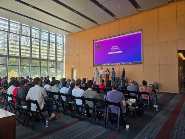 A photograph captures a presentation or seminar taking place in a modern auditorium with a large window wall, showing an audience seated in rows and a small group of five presenters standing near a large screen displaying the text 'Protect Your Brand.'
