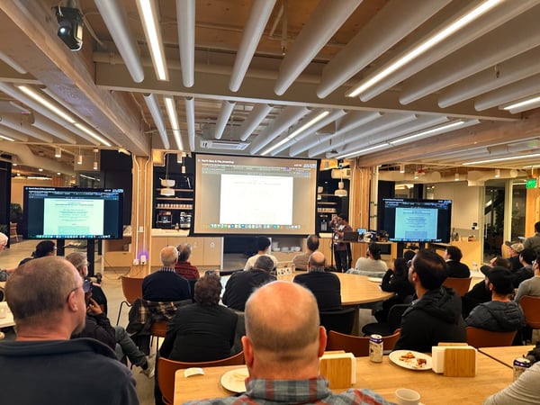 A photograph captures an indoor presentation or seminar in a modern event space, showing a speaker addressing a large audience seated at tables, with content displayed across a central projection screen and two flanking monitors.