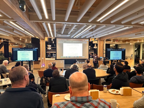 A photograph captures an indoor presentation or meeting where a speaker stands in front of a large projected screen and two side monitors, addressing a seated audience gathered around wooden tables.
