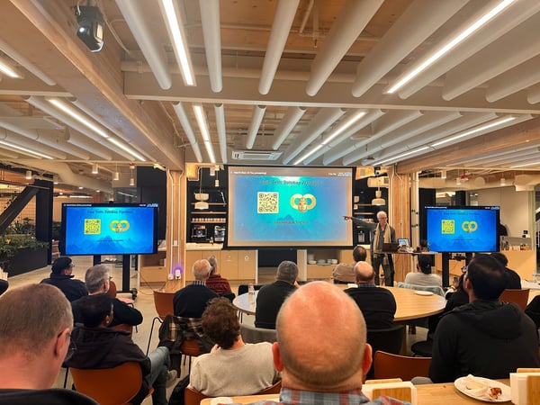 A photograph captures an indoor presentation taking place in a modern office space, featuring a speaker standing next to a large projected screen and a monitor displaying a slide titled "Key Learnings & Takeaways" to a seated audience.